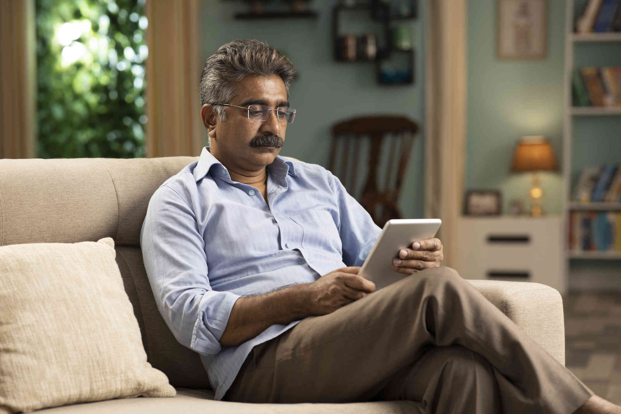 A middle aged man sits in an armchair in his home and looks down at the tablet in his hands with a serious expression.